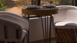 Una habitación con un gran ventanal, una mesa redonda de madera con un plato y una copa, dos sillas blancas y una planta en maceta.