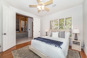 A bedroom with a bed, bedside lamps, a ceiling fan, a window with a view, and a door leading to another room.