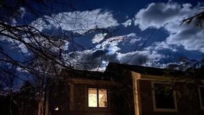 Kaksikerroksinen talo, jonka etuovi ja ikkunat ovat valaistuja, ja jota ympäröivät puut ja avoin, kirkas taivas.