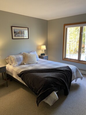 Een slaapkamer met een bed, nachtkastje, lamp en een raam met uitzicht op bomen.