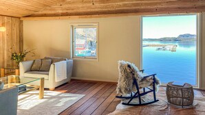 Une pièce chaleureuse avec un plafond en bois, une grande fenêtre donnant sur un lac et des bateaux, un canapé avec un plaid et un fauteuil à bascule.