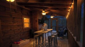 Interior d'una cabana de fusta amb una taula de dinar i tamborets, un ventilador de sostre i una finestra amb vistes a un bosc.