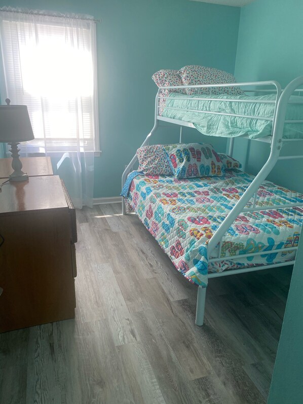 Een stapelbed met een kleurrijke sprei, een bureau met een lamp en een raam met dunne gordijnen.