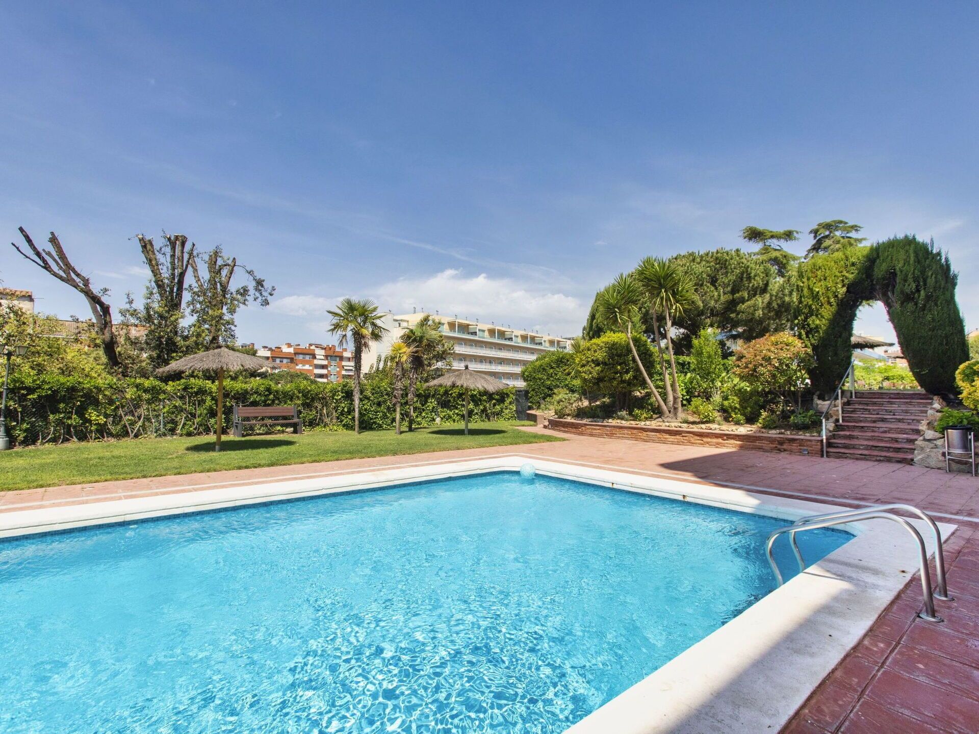 Una piscina rectangular con agua azul cristalina, rodeada por un patio de ladrillo, palmeras y un césped bien cuidado.