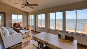A spacious living room with a large window overlooking a body of water, a ceiling fan, and a wooden coffee table.