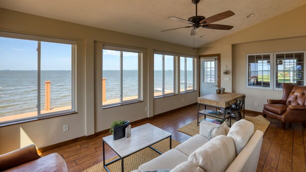 A spacious living room with large windows overlooking the sea, a ceiling fan, and a dining area.