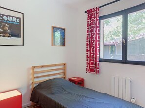 Une chambre à coucher avec un lit en bois, une table de chevet rouge, une affiche encadrée, une fenêtre avec un rideau rouge et blanc, et un radiateur.