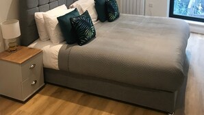 Ein modernes Schlafzimmer mit einem grau gepolsterten Bett, weißen und grünen Kissen, einem Nachttisch mit Lampe, einem gerahmten Kunstwerk an der Wand und Blick ins Grüne durch ein großes Fenster.