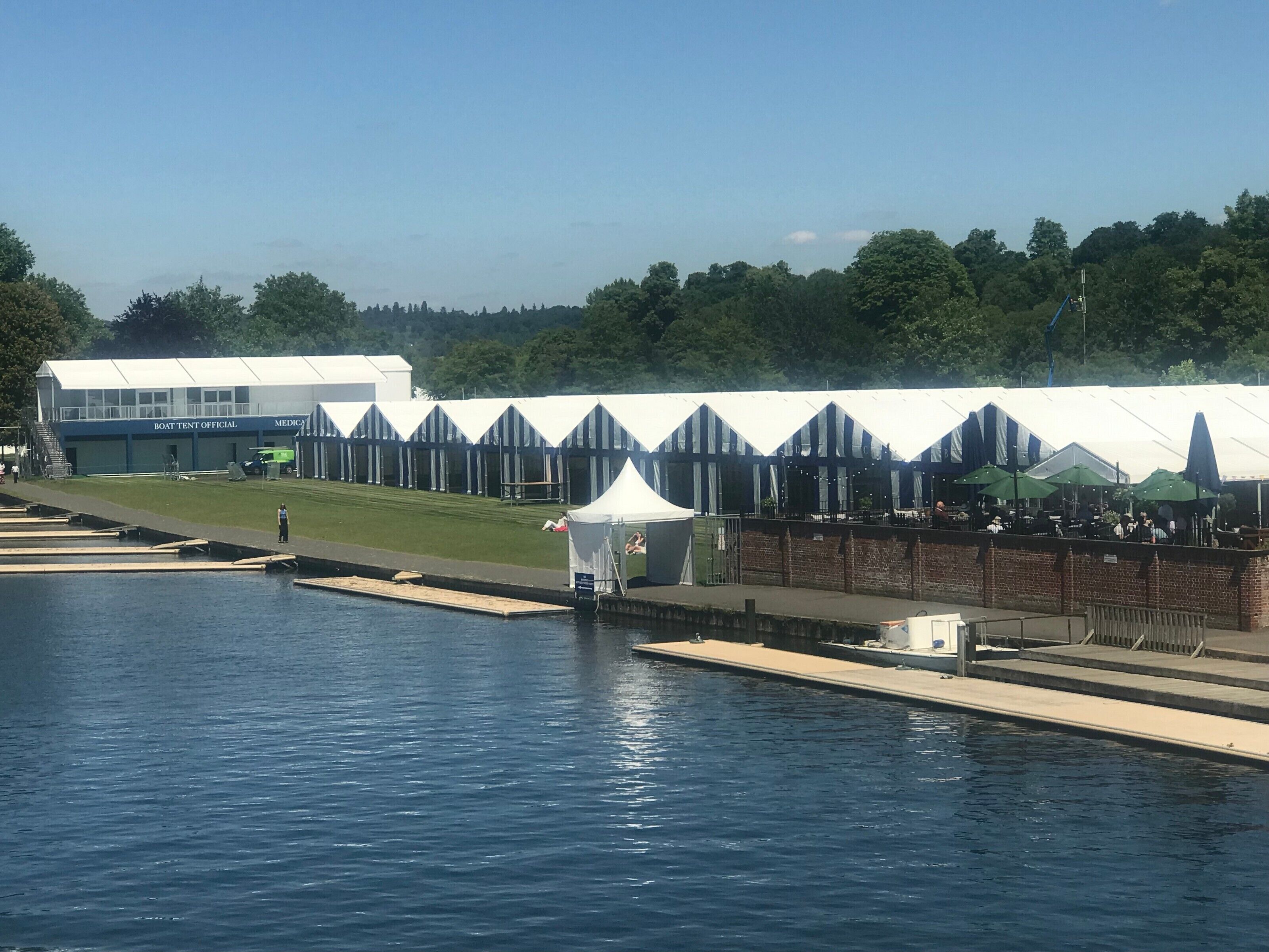A waterfront area with a boat tent official building, a grassy area, and a row of tents with tables and umbrellas.