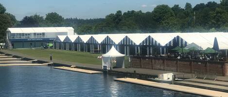 A waterfront area with a boat tent official building, a grassy area, and a row of tents with tables and umbrellas.