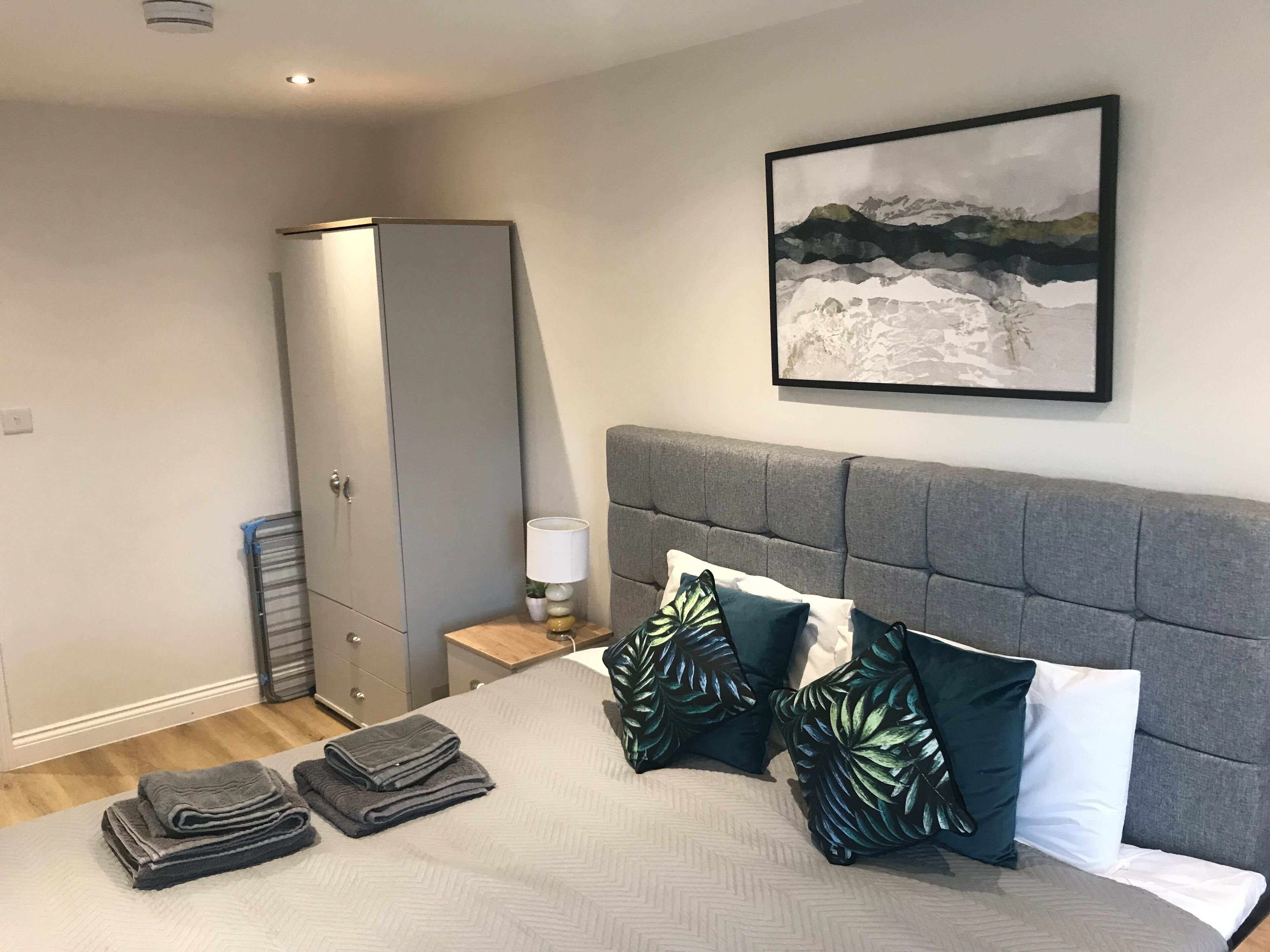 Un dormitorio con cama, cabecera gris, armario gris, mesita de noche con lámpara y un cuadro enmarcado en la pared.