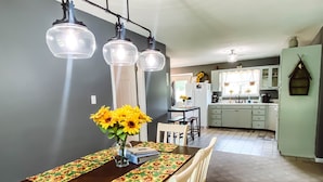 Een eetruimte met een lange tafel, stoelen en een vaas zonnebloemen. In de achtergrond is een keuken met kasten en een koelkast te zien.