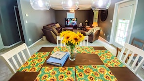 Een eetgedeelte met een tafel voor twee personen, een vaas met zonnebloemen en een televisie op de achtergrond.
