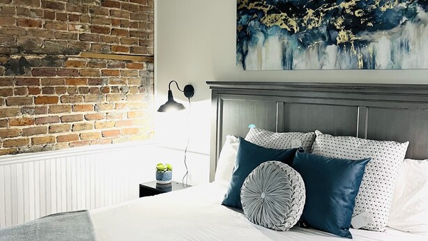 Un dormitorio con pared de ladrillo, un cuadro abstracto grande, una cama con ropa blanca y almohadas azules, y una mesita de noche con una lámpara.