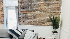 Una sala de estar con un sofá seccional gris, una alfombra blanco y negro, una pared de ladrillo, una planta en maceta y una pequeña mesa de madera.