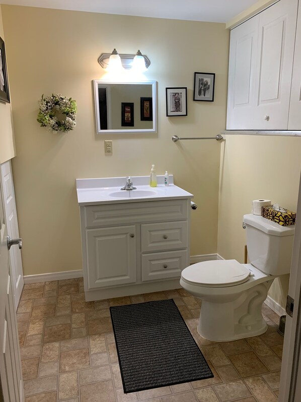 A bathroom with a white vanity, a toilet, a black doormat, and a wall-mounted mirror with lights.