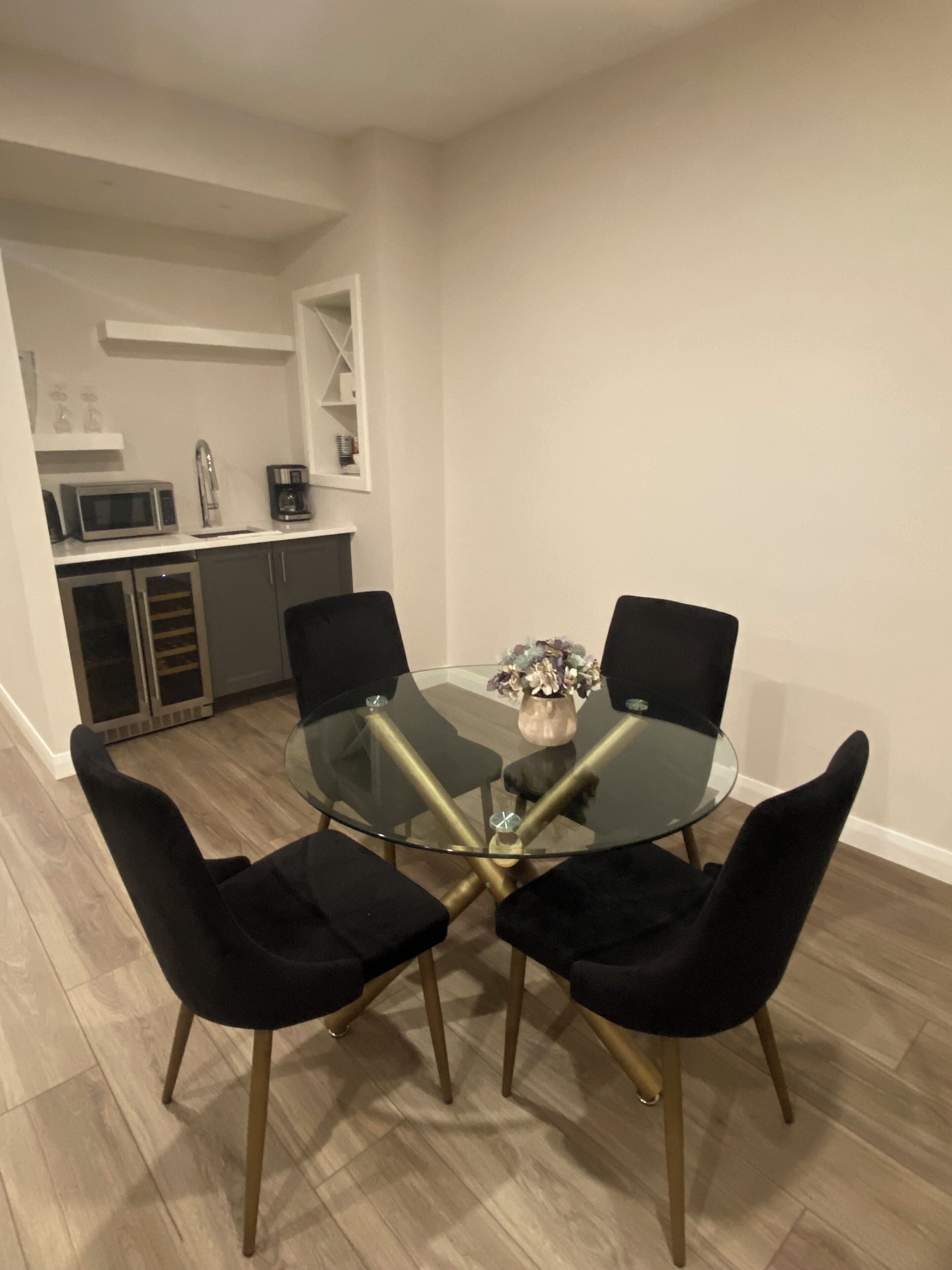 Un comedor con una mesa de vidrio rodeada por cuatro sillas negras con patas doradas. Hay un jarrón pequeño con flores sobre la mesa.