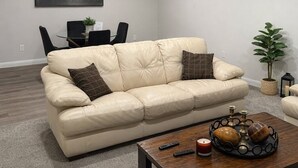 En moderne stue med en beige lædersofa, et træbord med stearinlys og et spiseområde med et glasbord og stole.