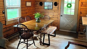 A wooden cabin interior with a dining table, chairs, a bench, and a door with a wreath.