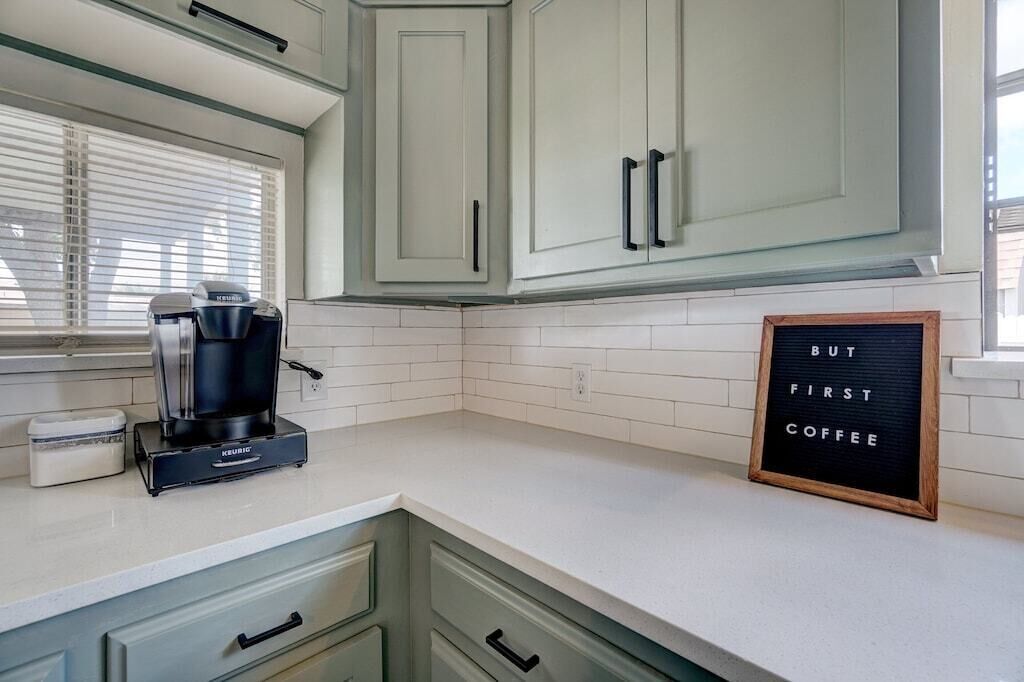 Una cocina con gabinetes blancos, una cafetera y un cartel enmarcado que dice "BUT FIRST COFFEE".