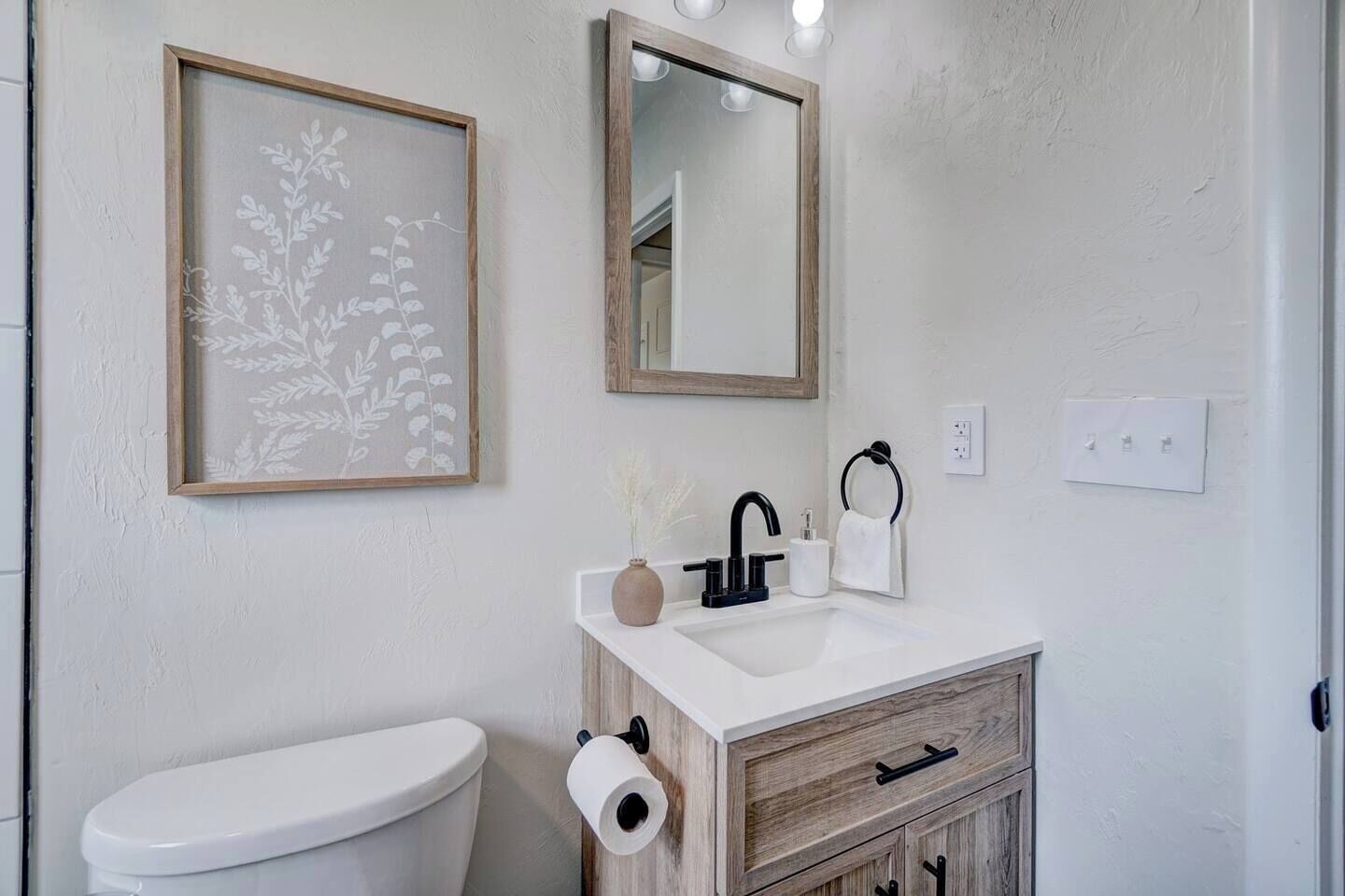 Ein Badezimmer mit Toilette, Waschbecken mit Spiegel, einer gerahmten Pflanze und einem Handtuchhalter.