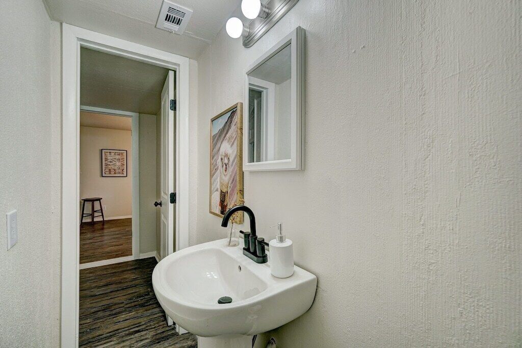 A bathroom with a white sink, black faucet, and a mirror above it. There is a framed picture on the wall and a stool in the adjacent room.