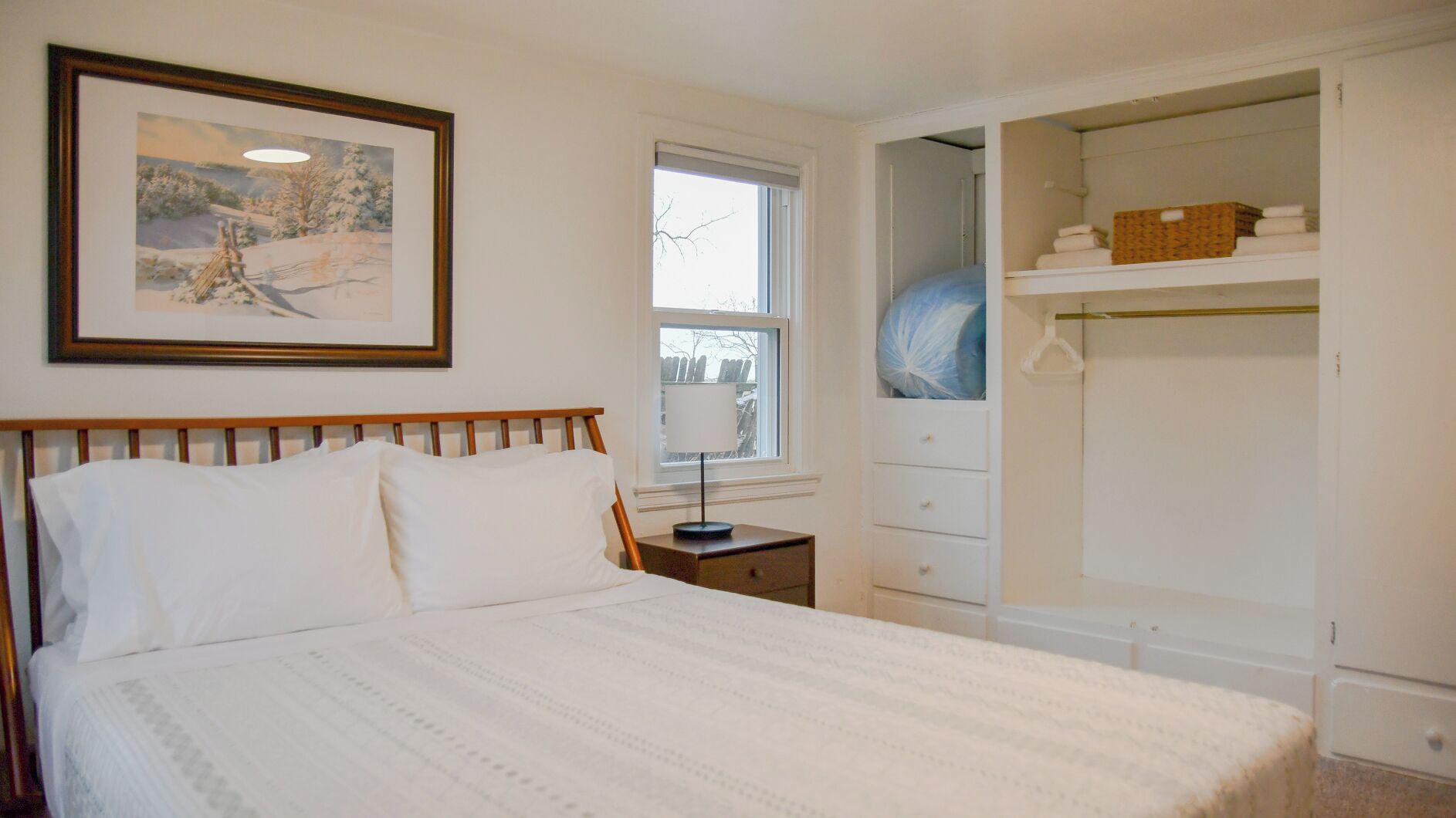 A bedroom with a bed, a nightstand, a window, a framed picture, and a closet with shelves.