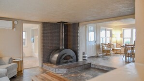 Un salon chaleureux avec une cheminée, un coin repas avec des chaises en osier et une cuisine visible à travers une porte ouverte.