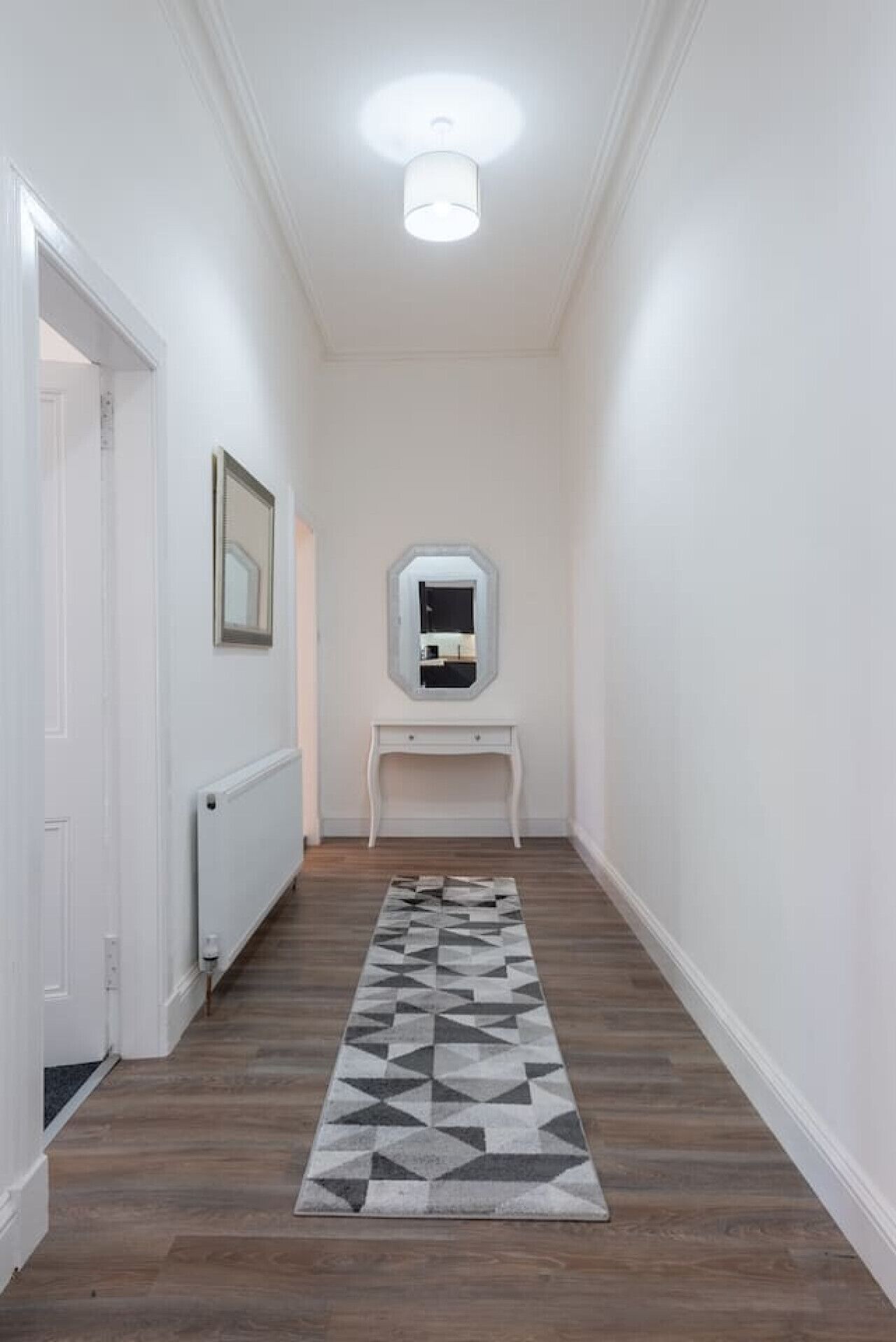 Un couloir étroit avec un tapis à motifs, une console blanche surmontée d’un miroir et un radiateur fixé au mur.