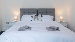 Een net opgemaakt bed met witte lakens, een grijze hoofdbord en twee bedlampjes.