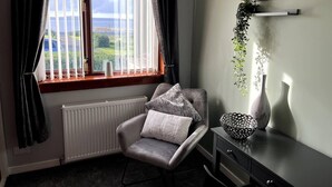 Una habitación con vistas a través de una ventana, un sillón gris con un cojín, un radiador, una mesita con un cuenco y un jarrón, y una estantería con una planta y un letrero.