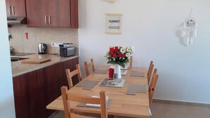 En matplats med ett träbord, stolar, en vas med blommor och ett köksområde med skåp och köksmaskiner.