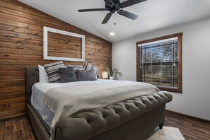 Un dormitorio con pared de madera, una cama con cabecera capitonada, un ventilador de techo y una ventana con persianas.