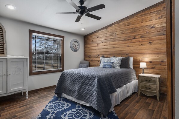 Un dormitorio con pared de madera, un ventilador de techo, una cama con colcha azul y blanca, una mesita de noche con lámpara y una ventana con persianas.