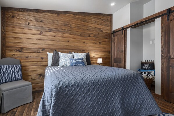 Un dormitorio con cabecera de madera, una cama con colcha azul, un sillón gris y una mesita de noche con lámpara.
