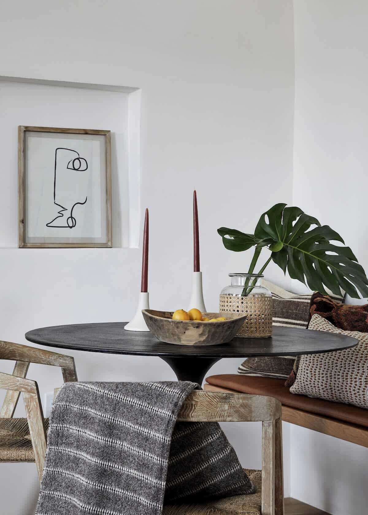 A round table with a bowl of fruit, two candles, and a framed abstract art piece on the wall.