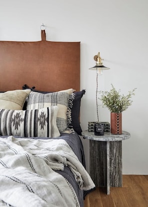 Een slaapkamer met een bed, kussens, een nachtkastje met een lamp en een vaas met bloemen.