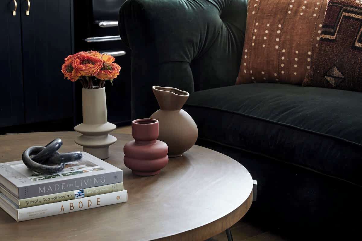 A round wooden coffee table with a stack of books, a vase with orange flowers, and two ceramic vases.