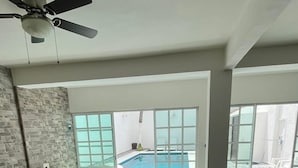 A spacious living room with a beige sectional sofa, a black coffee table, and a ceiling fan. Large windows provide a view of an outdoor swimming pool. - Magnífica casa con piscina privada a 10 min del mar (Cancún)