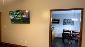 Ein Wohnzimmer mit Wand-Fernseher, einem Essbereich mit Tisch und Stühlen sowie einem Flur, der zu einer Küchenzeile führt.