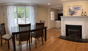 A dining area with a wooden table and chairs, a fireplace with a mantelpiece, and a view of the outdoors through a window with curtains.