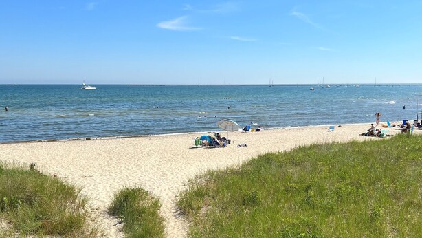 Ein Strand mit sandigem Ufer, ein paar Boote im Wasser und Menschen, die unter Sonnenschirmen entspannen.
