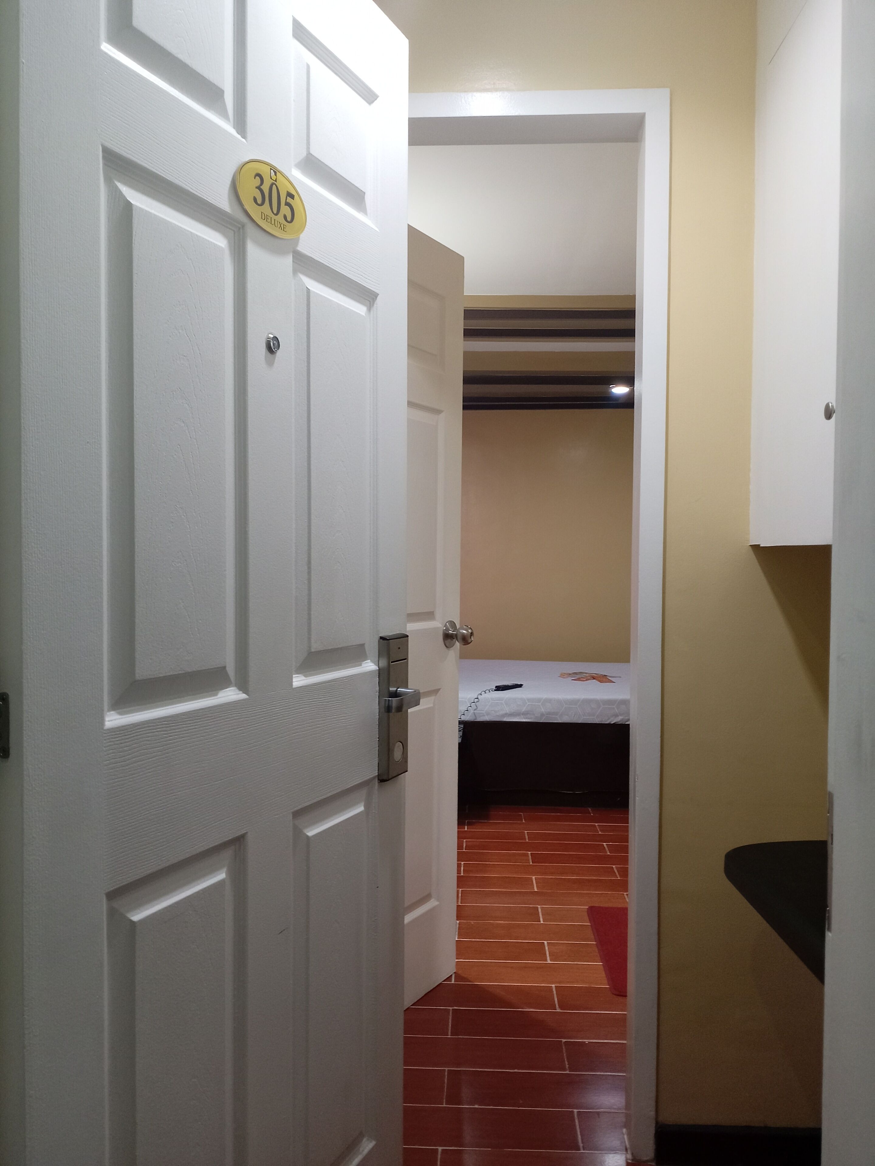Una habitación de hotel con una puerta blanca que indica "305 Deluxe", una cama con cubrecama blanco y una alfombra roja en el suelo.