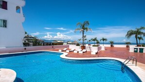 Una piscina con sillas de descanso y palmeras, rodeada por un edificio blanco con vista al océano.