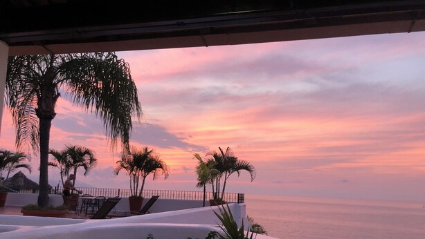 Terraza con vista al mar al atardecer, con palmeras y mobiliario exterior.