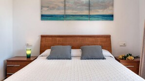 A bedroom with a wooden headboard, a bed with white bedding, two gray pillows, a nightstand with a lamp, and a wall-mounted painting of a seascape.