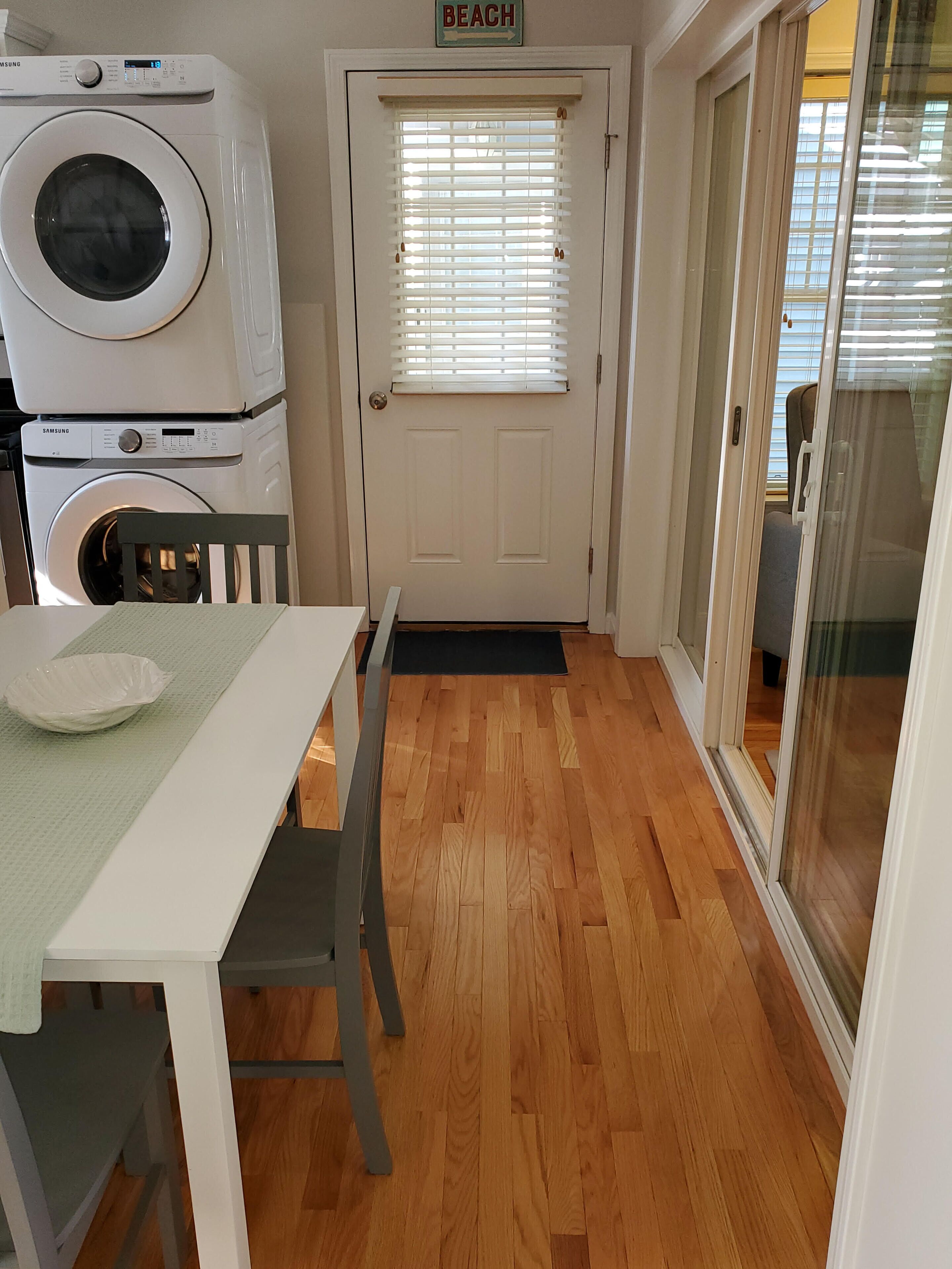 Un espace buanderie avec des machines à laver et des sèche-linge superposés, une porte sur laquelle est affiché le mot « PLAGE », et une table avec des chaises.