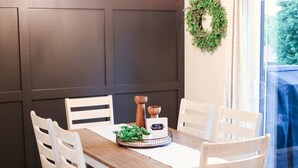 Uma sala de jantar com uma mesa de madeira, cadeiras brancas, painéis de parede escuros, um candelabro e vista através de uma porta de vidro.
