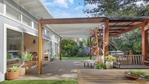 Un patio cubierto con una mesa y sillas de madera, rodeado de plantas en macetas y un césped.