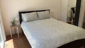 Una cama bien hecha con una colcha estampada, una mesita de noche con una lámpara y una planta, y una silla en la esquina.